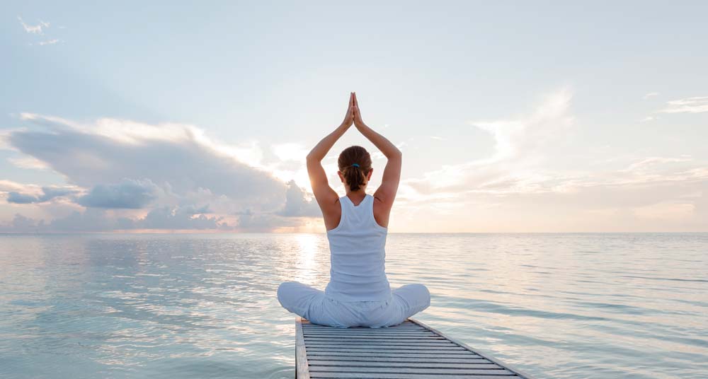 Frau sitzt im Yoga-Sitz mit Blick aufs Meer entspannt auf einem Steg