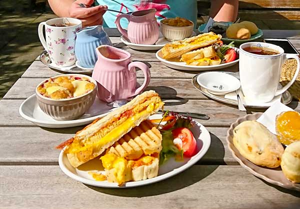 Reich gedeckter Tisch mit Sandwiches, Crumbles, Scones, Tee, Kaffee im Sonnenschein des Gartens