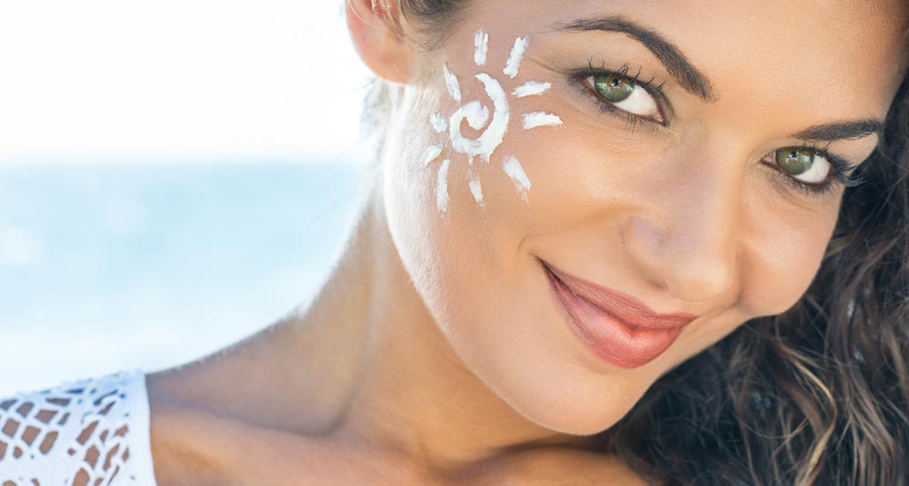 Frau mit einer Sonne aus Sonnencreme auf der Wange am Strand lächelt in die Kamera