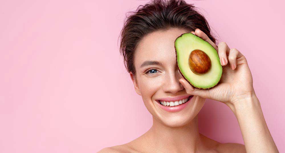Junge Frau hält sich eine halbierte Avocado mit Kern vor ihr linkes Auge und lächelt