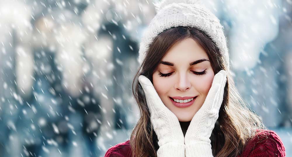 Lächelnde Frau mit Wollmütze steht mit geschlossenen Augen im Schneegestöber, trägt Handschuhe und hält sich die Hände an die Wangen