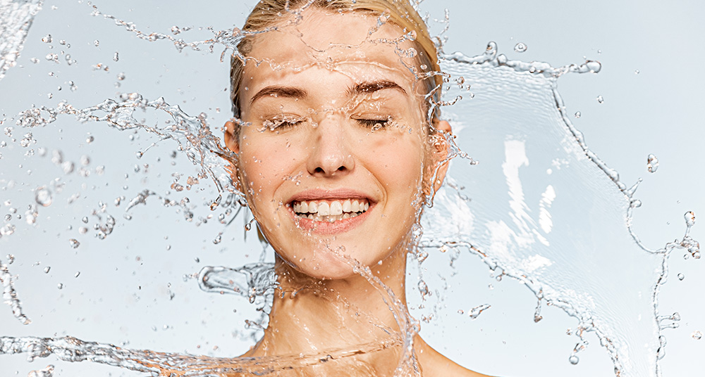 Foto von jungen Frauen mit sauberer Haut und Wassersplash. Portraet einer laechelnden Frau mit Tropfen Wasser um ihr Gesicht.